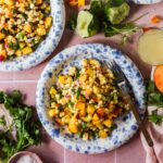 A plate of corn and peach salad with diced red onion and herbs, surrounded by cilantro, lime halves, and a fork on a pink surface.