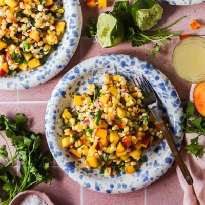 A plate of corn and peach salad with diced red onion and herbs, surrounded by cilantro, lime halves, and a fork on a pink surface.