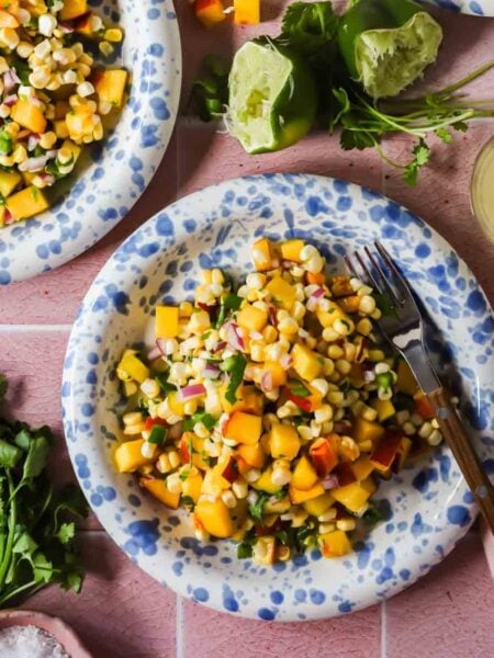 A plate of corn and peach salad with diced red onion and herbs, surrounded by cilantro, lime halves, and a fork on a pink surface.
