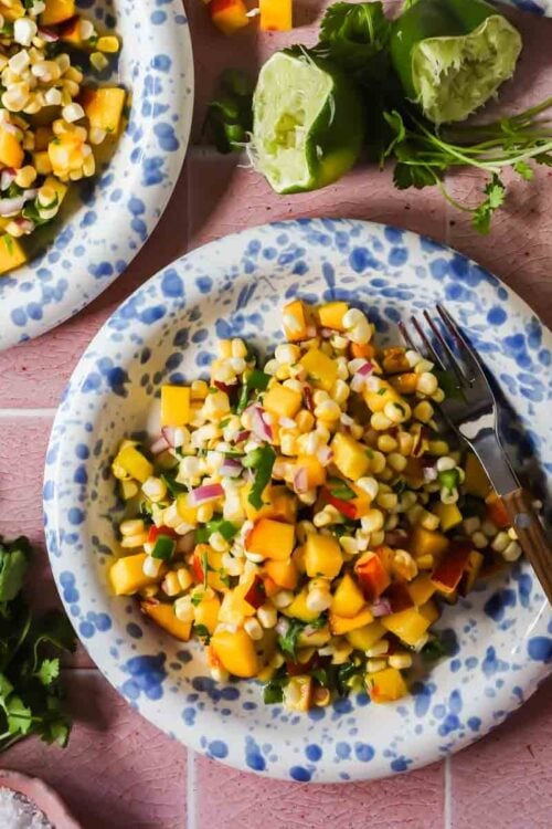 A plate of corn and peach salad with diced red onion and herbs, surrounded by cilantro, lime halves, and a fork on a pink surface.