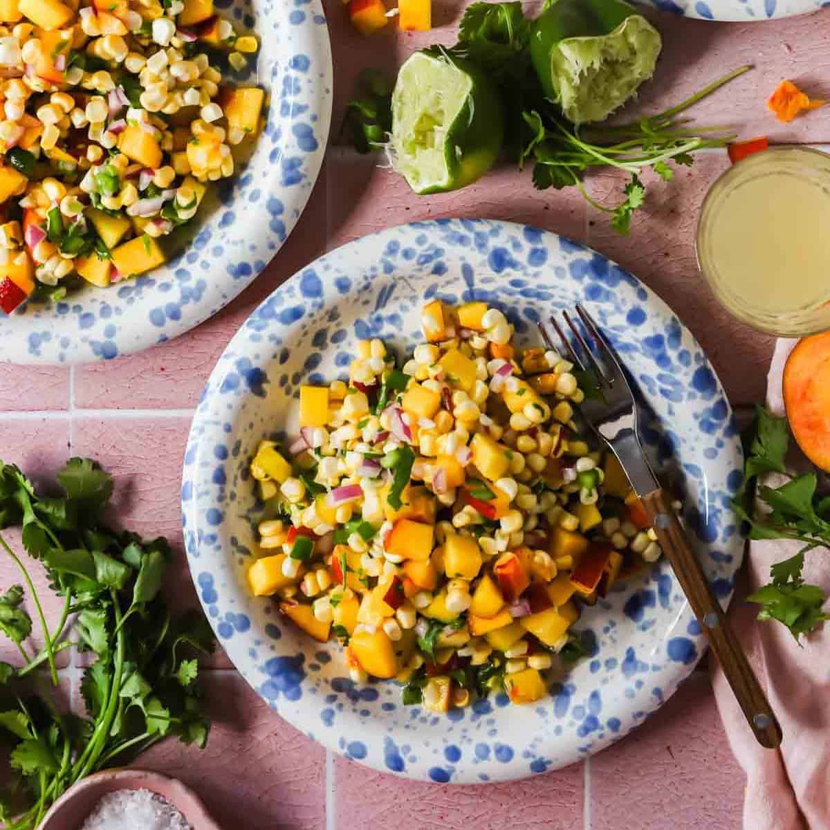 A plate of corn and peach salad with diced red onion and herbs, surrounded by cilantro, lime halves, and a fork on a pink surface.