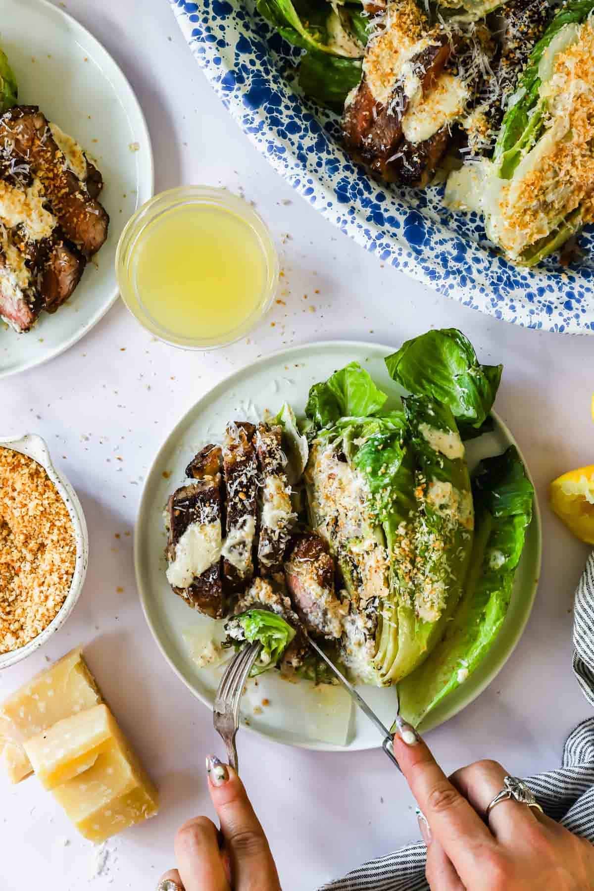 A person uses a knife and fork to cut into grilled lettuce and steak on a plate, topped with grated cheese, next to a bowl of breadcrumbs and a glass of dressing.