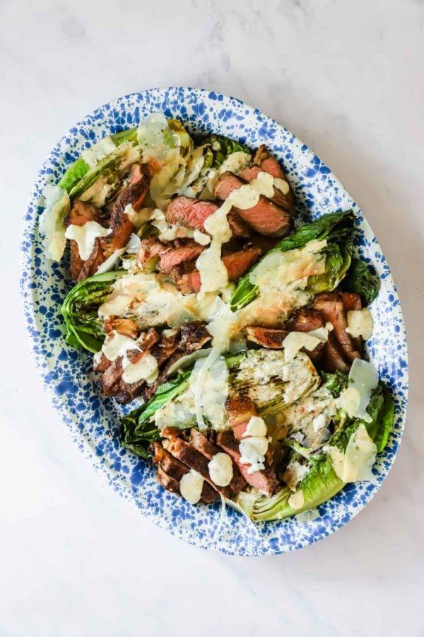 A platter with grilled steak slices, charred romaine lettuce, shaved parmesan, and creamy dressing on a blue and white speckled dish.