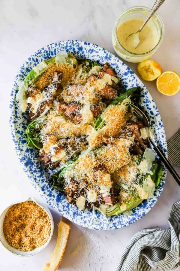 A platter of grilled romaine lettuce topped with grated cheese, croutons, and creamy dressing, with lemon halves and a jar of dressing nearby.