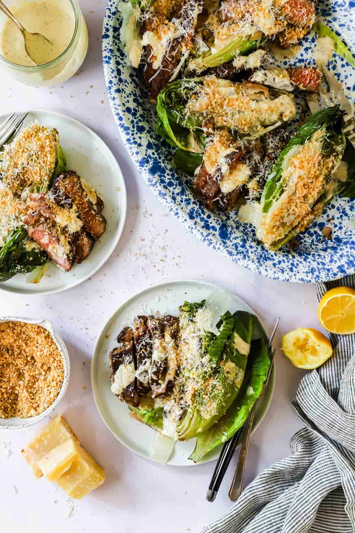 Plates and a serving dish with grilled romaine lettuce, steak slices, grated cheese, breadcrumbs, and dressing, garnished with lemon halves and a wedge of parmesan nearby.