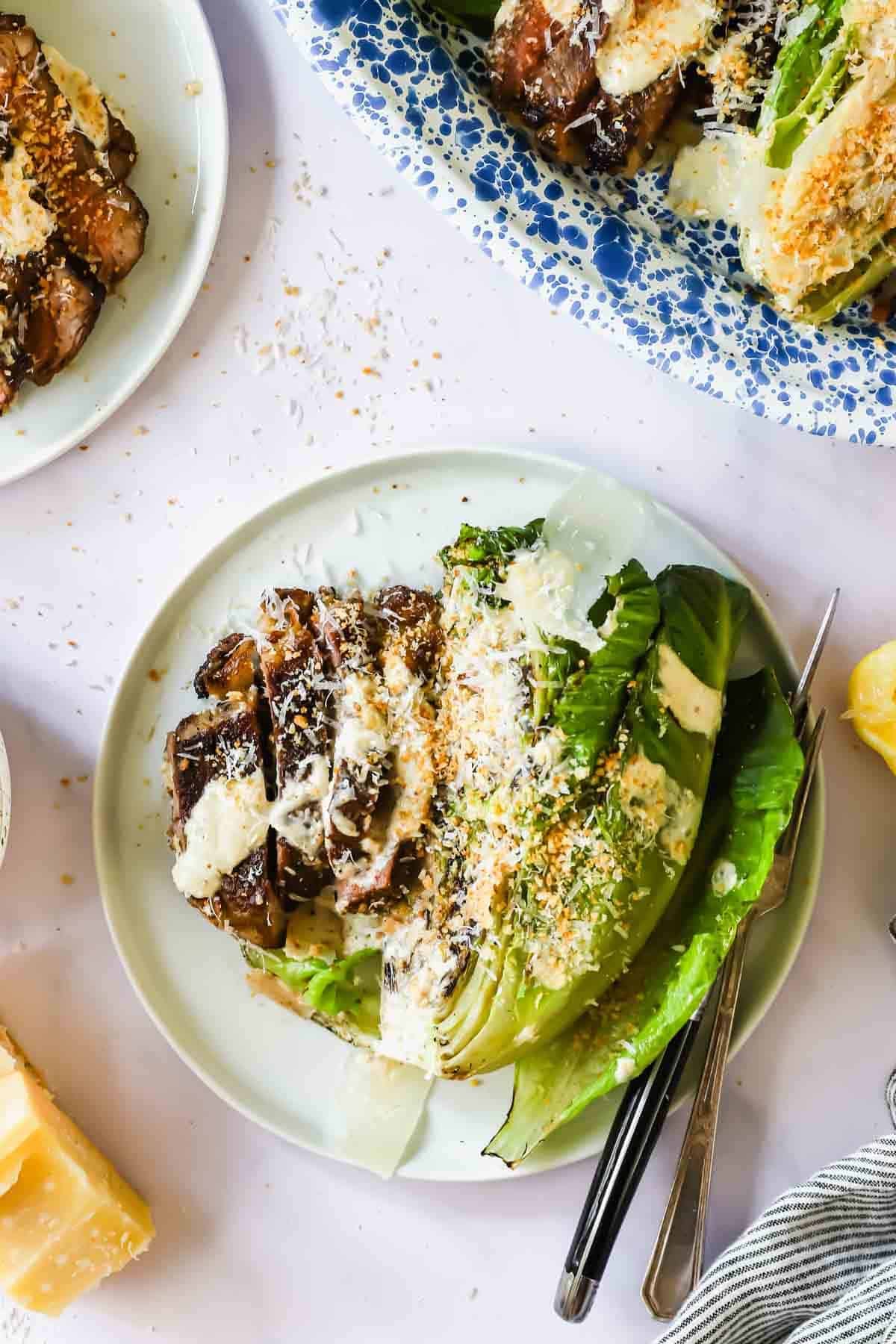 A plate with grilled romaine lettuce, sliced steak, grated cheese, and creamy dressing, served with a fork and knife.