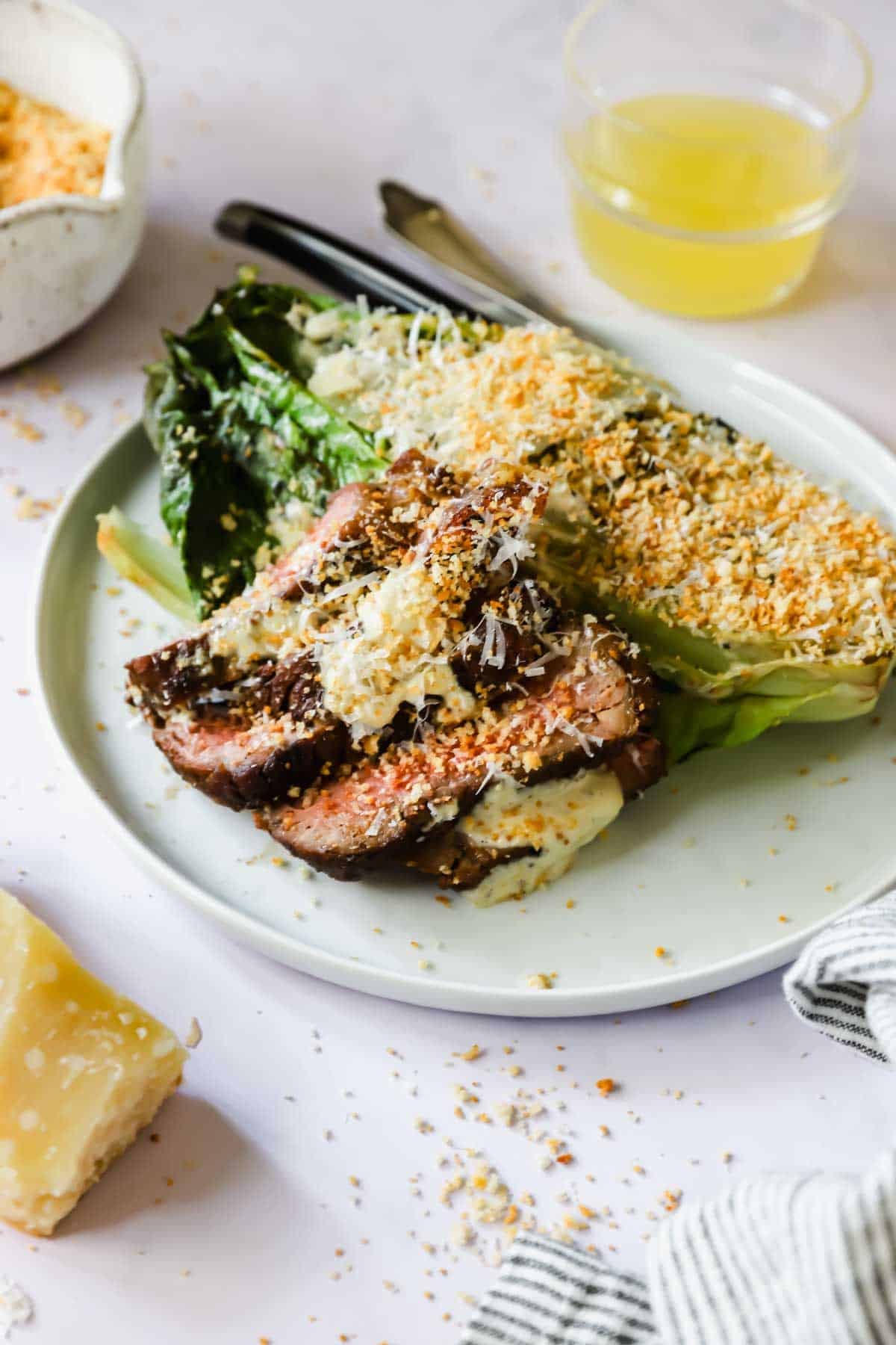 A plate with sliced grilled steak, charred romaine lettuce topped with grated cheese and breadcrumbs, and a glass of yellow liquid in the background.
