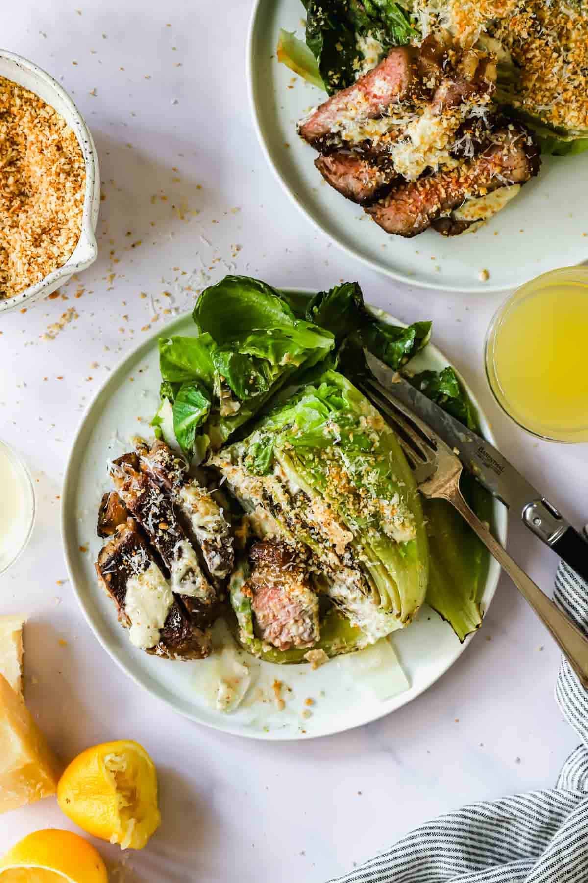 A plate with grilled romaine lettuce, sliced steak, grated cheese, and toasted breadcrumbs, next to a glass of juice and lemon halves.