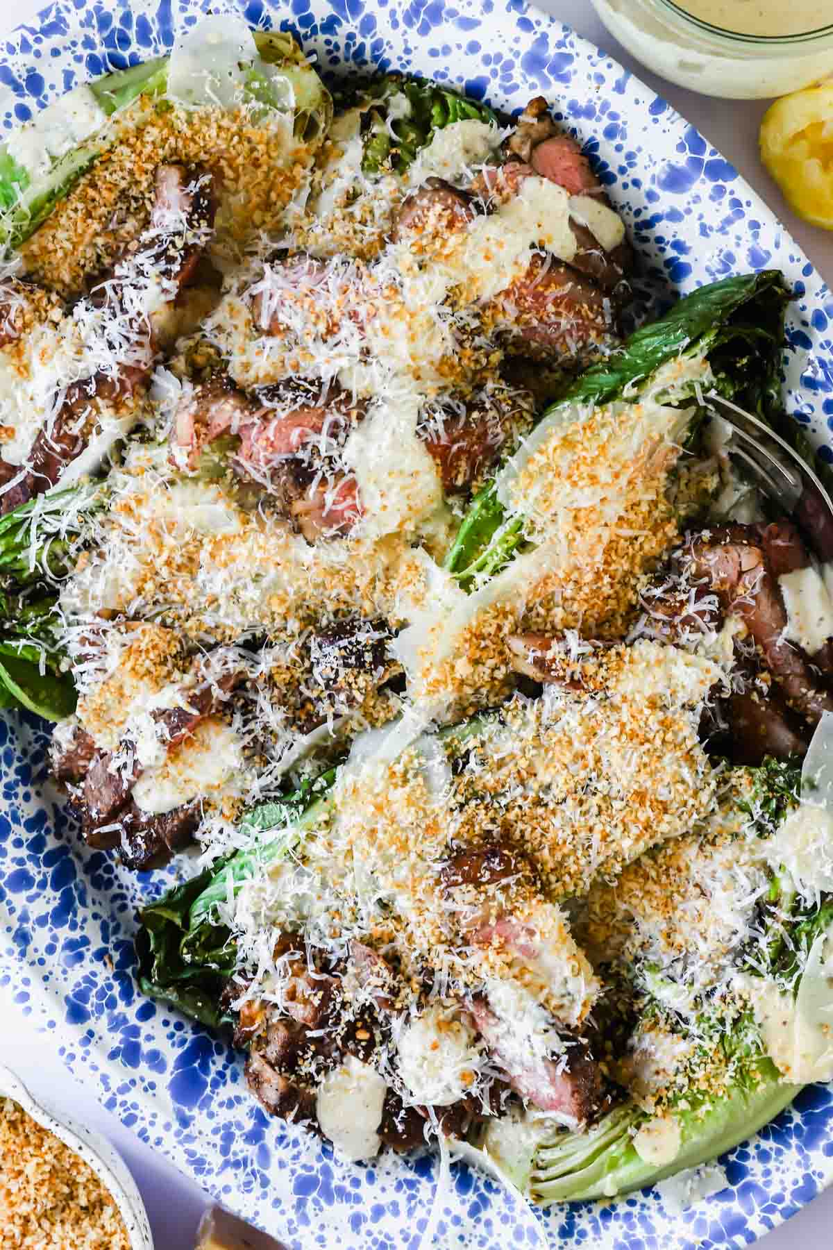 A platter of grilled romaine lettuce topped with sliced steak, grated cheese, toasted breadcrumbs, and creamy dressing on a blue and white patterned plate.