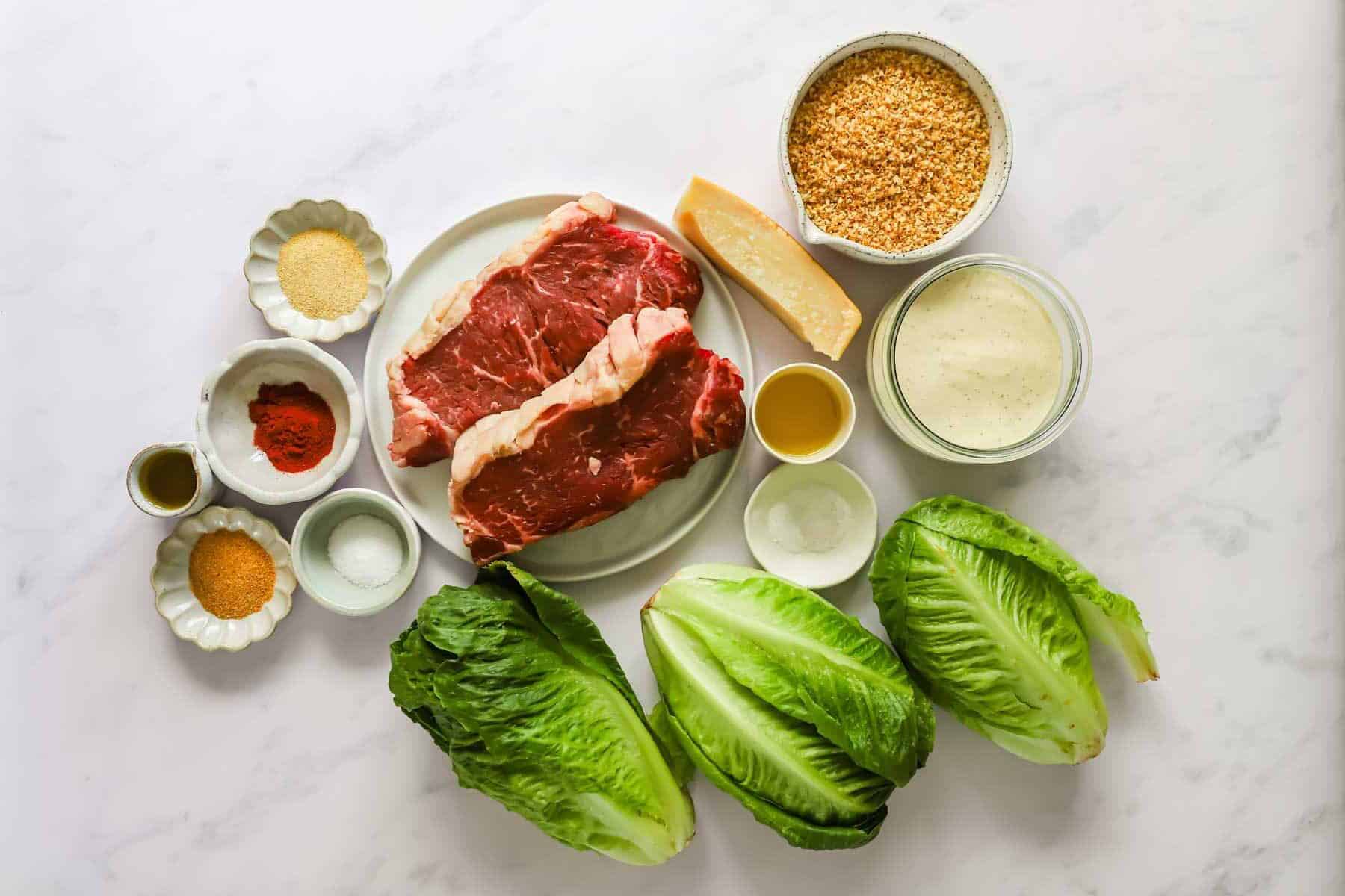 Two raw steaks on a plate surrounded by romaine lettuce, grated cheese, breadcrumbs, Caesar dressing, and small bowls of seasonings and olive oil on a white surface.