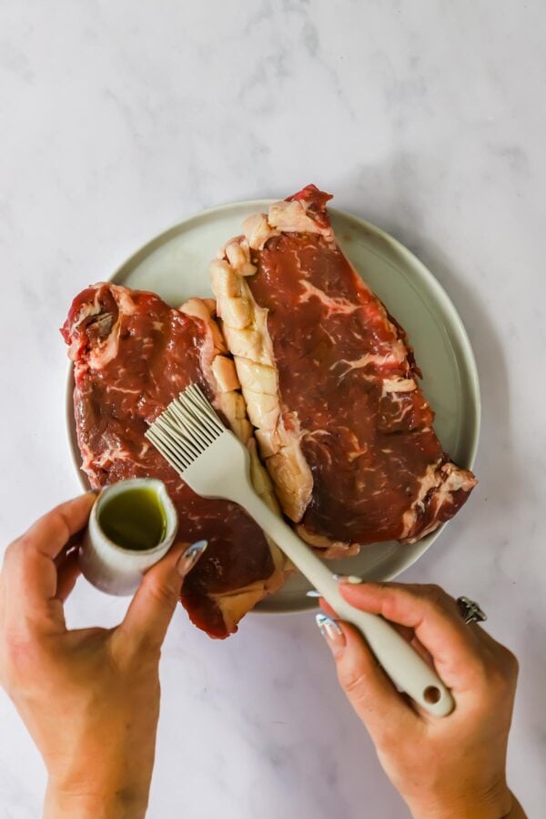 Two raw steaks on a plate being brushed with oil; a hand holds a small container of oil and another hand uses a brush to apply it to the meat.