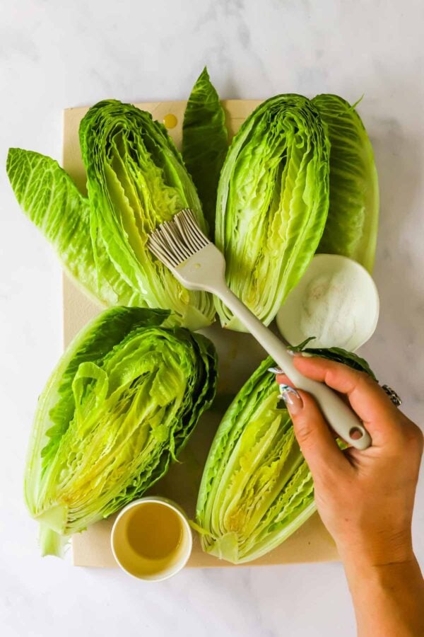 A hand uses a brush to coat halved romaine lettuce heads on a cutting board, with small bowls of oil and salt nearby.