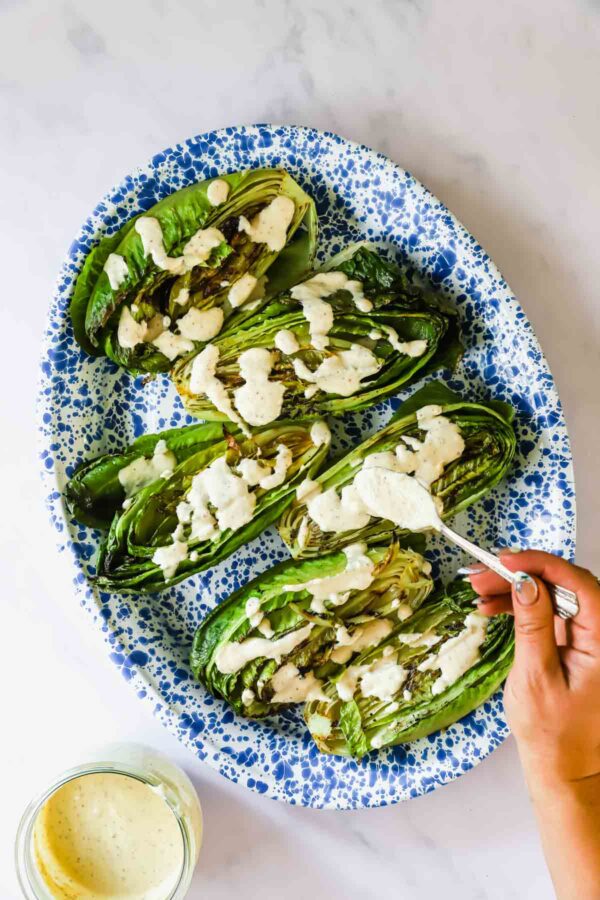 A hand drizzles creamy dressing over grilled romaine lettuce halves arranged on a blue and white speckled oval platter.