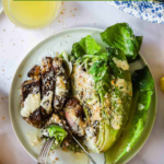 A plate with steak Caesar salad topped with homemade dressing, accompanied by a glass of drink; person cutting into steak with a fork and knife.