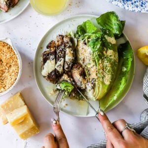 A person slices into grilled romaine lettuce with creamy dressing and breadcrumbs on a plate, with Parmesan cheese and a bowl of extra breadcrumbs beside it.