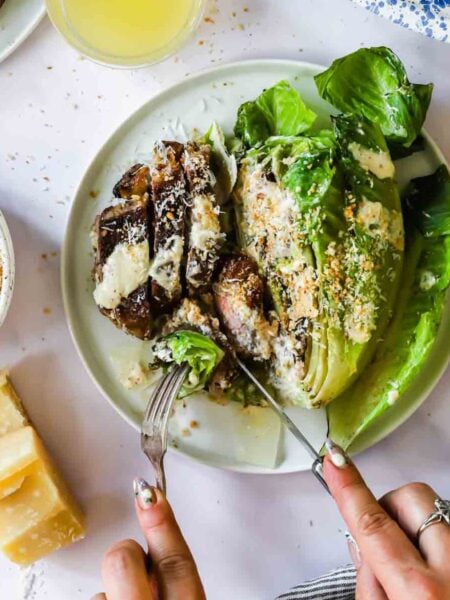 A person slices into grilled romaine lettuce with creamy dressing and breadcrumbs on a plate, with Parmesan cheese and a bowl of extra breadcrumbs beside it.