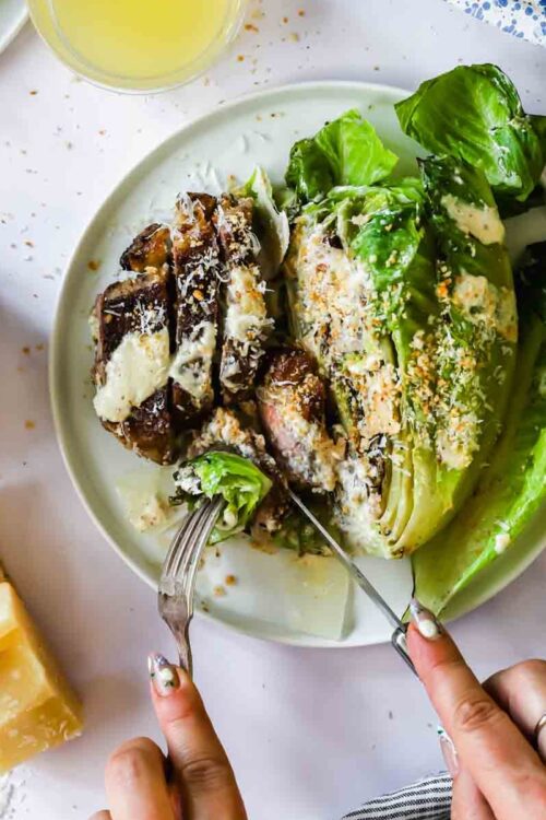 A person slices into grilled romaine lettuce with creamy dressing and breadcrumbs on a plate, with Parmesan cheese and a bowl of extra breadcrumbs beside it.