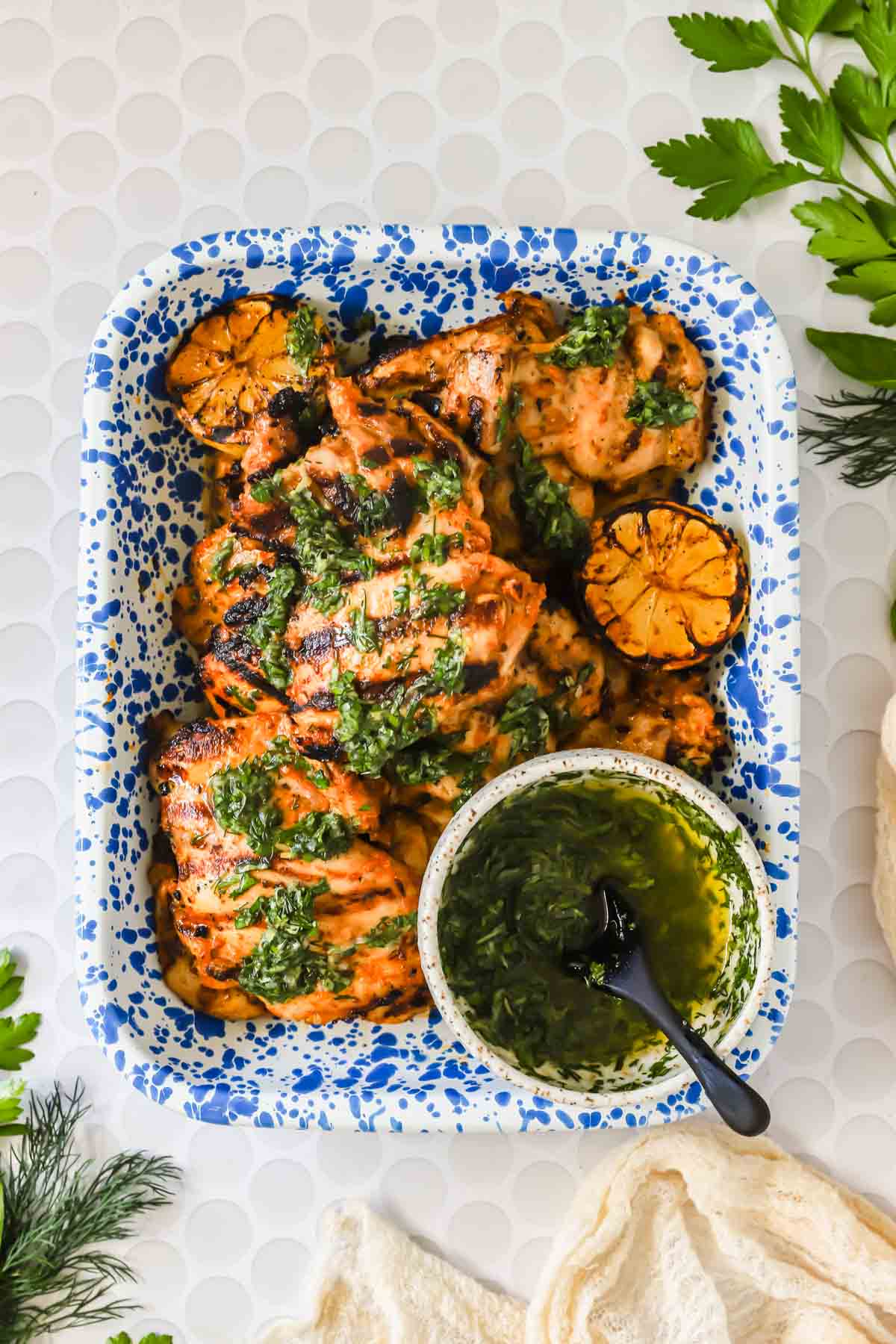 Grilled chicken pieces topped with green herb sauce, served with charred lemon halves on a blue and white tray, next to a bowl of sauce with a spoon.