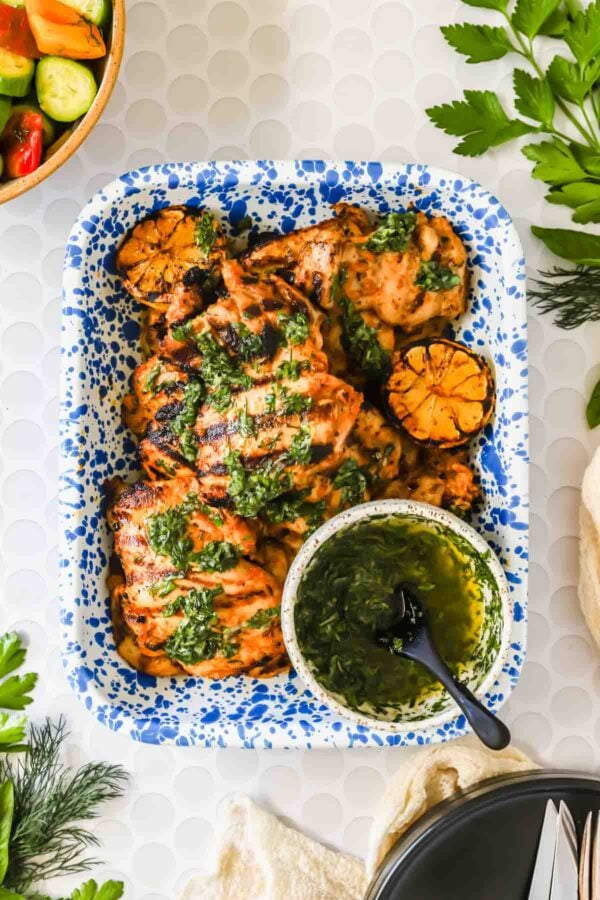 A blue and white tray with grilled chicken thighs, charred lemon halves, and a bowl of green herb sauce with a spoon, surrounded by herbs and salad.