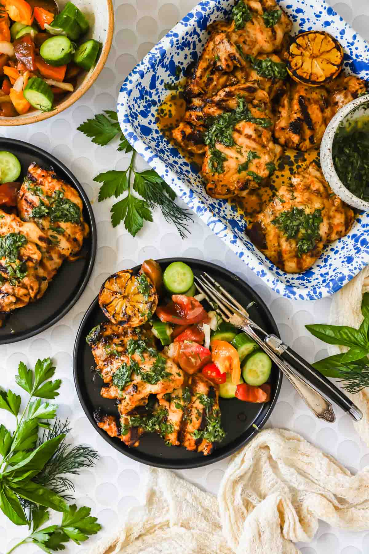 Grilled chicken topped with green herb sauce, served with a cucumber and tomato salad on black plates, with extra chicken and lemon halves in a blue-and-white dish.