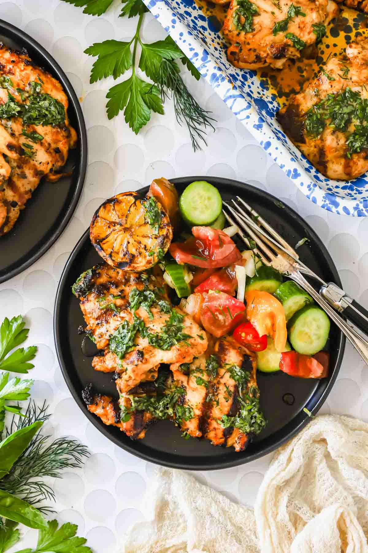 Plate with grilled chicken, charred lemon, cucumber-tomato salad, and herbs. Two forks rest on the plate. Additional chicken and herbs appear nearby.