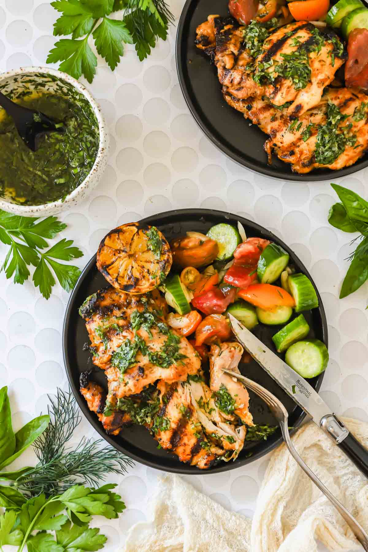 Two plates with grilled chicken topped with herbs, a grilled lemon half, and a fresh salad with cucumbers and tomatoes; a bowl of green sauce and fresh herbs are nearby.
