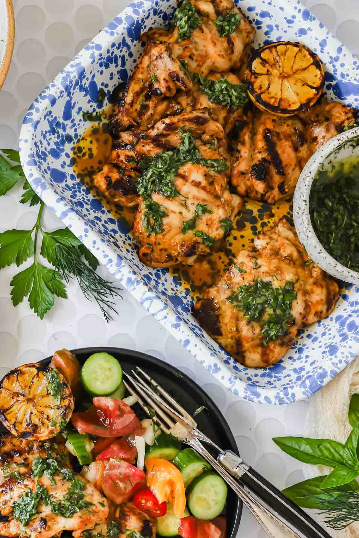 Grilled chicken thighs with chimichurri sauce in a blue and white dish, served with grilled lemon halves and a side plate of cucumber and tomato salad.