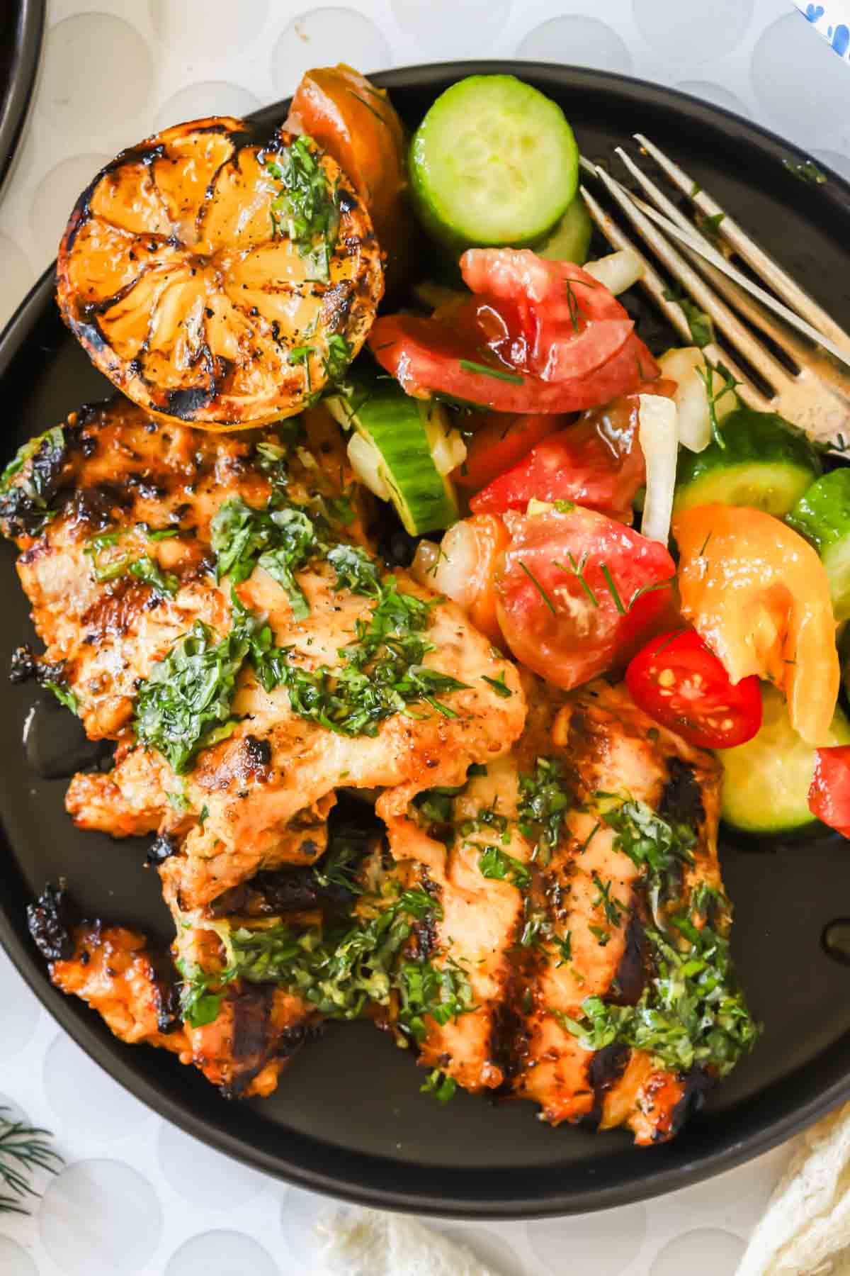Grilled chicken thighs topped with herbs, served with a mixed salad, sliced cucumber, and a halved grilled lemon on a black plate.
