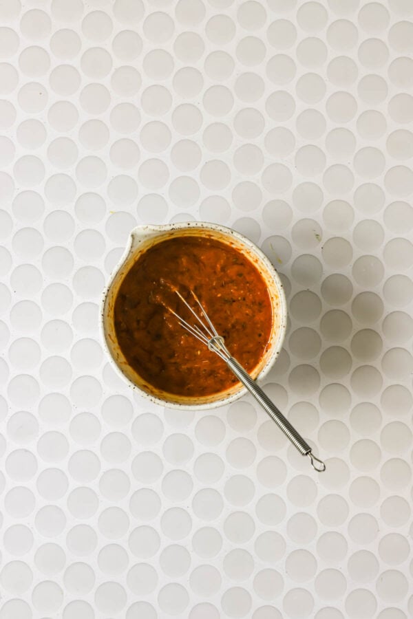 A small whisk rests in a bowl filled with reddish-brown sauce, placed on a white, circular-tiled surface.
