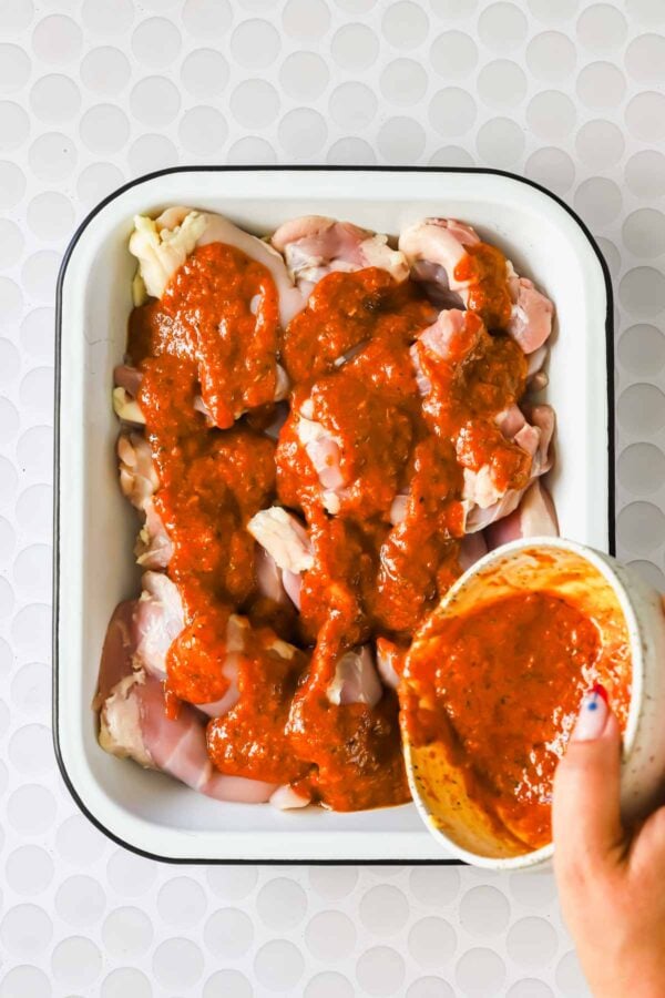 Raw chicken pieces in a white tray are being covered with red sauce poured from a bowl by a hand.