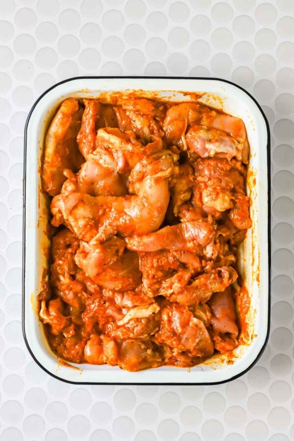Raw chicken pieces coated in a red marinade are arranged in a white rectangular dish on a white textured surface.