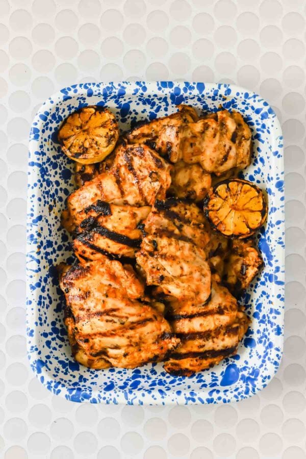 Blue speckled tray with grilled chicken thighs and charred lemon halves on a white textured surface.