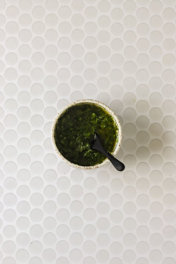 A bowl of green sauce with a black spoon sits on a white, circular-tiled surface.