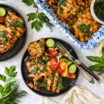 Grilled chicken with green herb sauce on black plates, served with sliced cucumbers, tomatoes, and charred lemon, surrounded by fresh herbs.