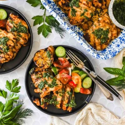 Grilled chicken with green herb sauce on black plates, served with sliced cucumbers, tomatoes, and charred lemon, surrounded by fresh herbs.