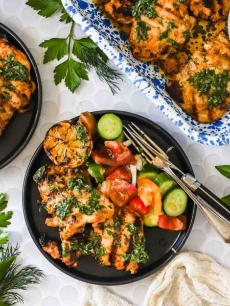 Grilled chicken with green herb sauce on black plates, served with sliced cucumbers, tomatoes, and charred lemon, surrounded by fresh herbs.