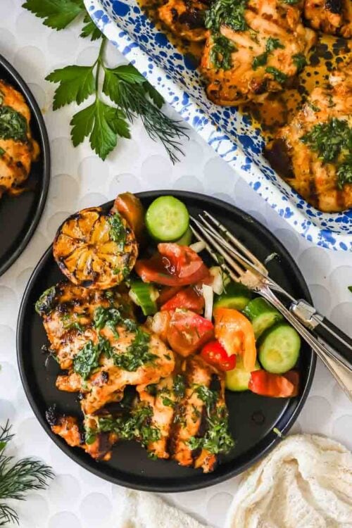 Grilled chicken with green herb sauce on black plates, served with sliced cucumbers, tomatoes, and charred lemon, surrounded by fresh herbs.