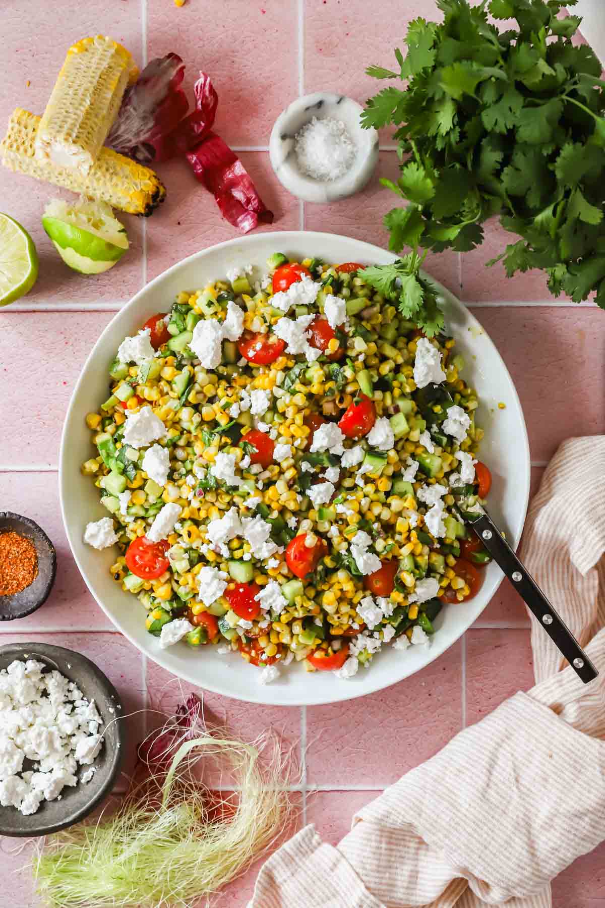 A bowl of corn salad with cherry tomatoes, avocado, cilantro, and crumbled cheese sits on a pink tile surface, surrounded by salad ingredients and seasonings.