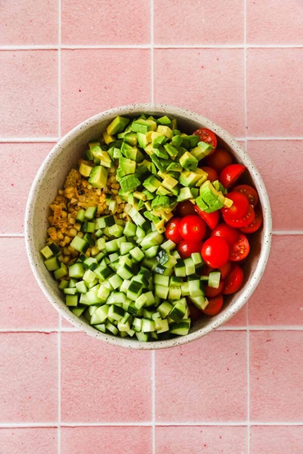 A bowl containing diced avocado, cherry tomatoes, diced cucumber, and corn sits on a pink tiled surface.