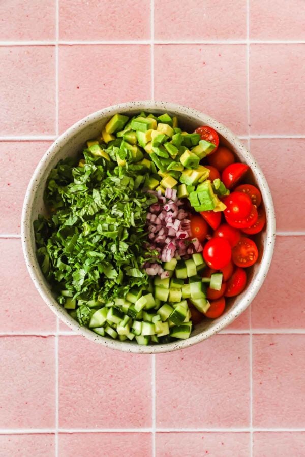 Bowl of chopped cucumber, cherry tomatoes, red onion, avocado, and cilantro on a pink tiled surface.