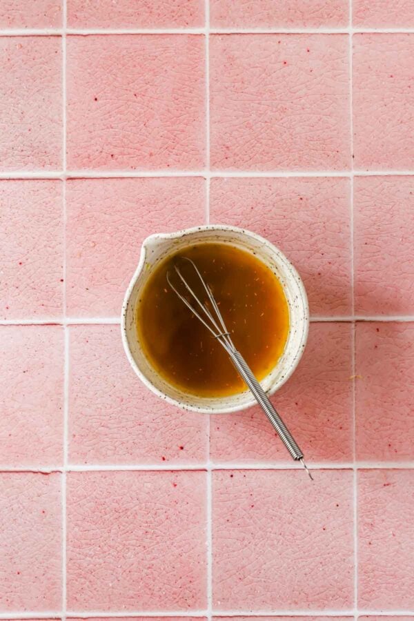 A small bowl of brown liquid with a metal whisk rests on pink tiled surface, viewed from above.