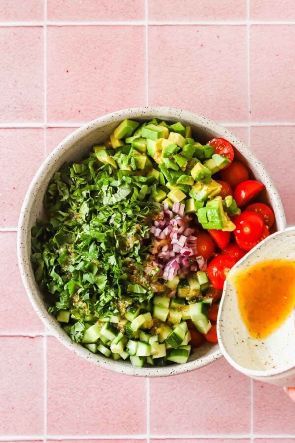 A bowl with chopped cucumber, avocado, tomato, onion, and cilantro with dressing being poured in, against a pink tiled background.