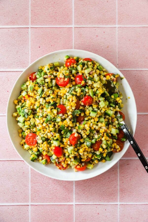 Bowl of corn salad with cherry tomatoes, cucumber, herbs, and a spoon, placed on a pink tiled surface.