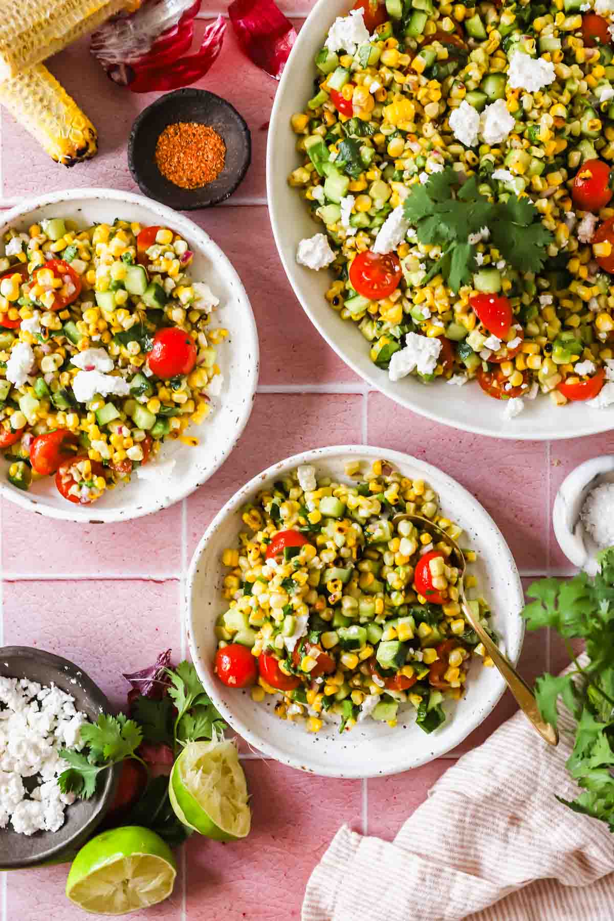 Three bowls of corn salad with tomatoes, cucumber, cilantro, and cheese are arranged on a pink tiled surface, surrounded by lime wedges, seasoning, and salad ingredients.