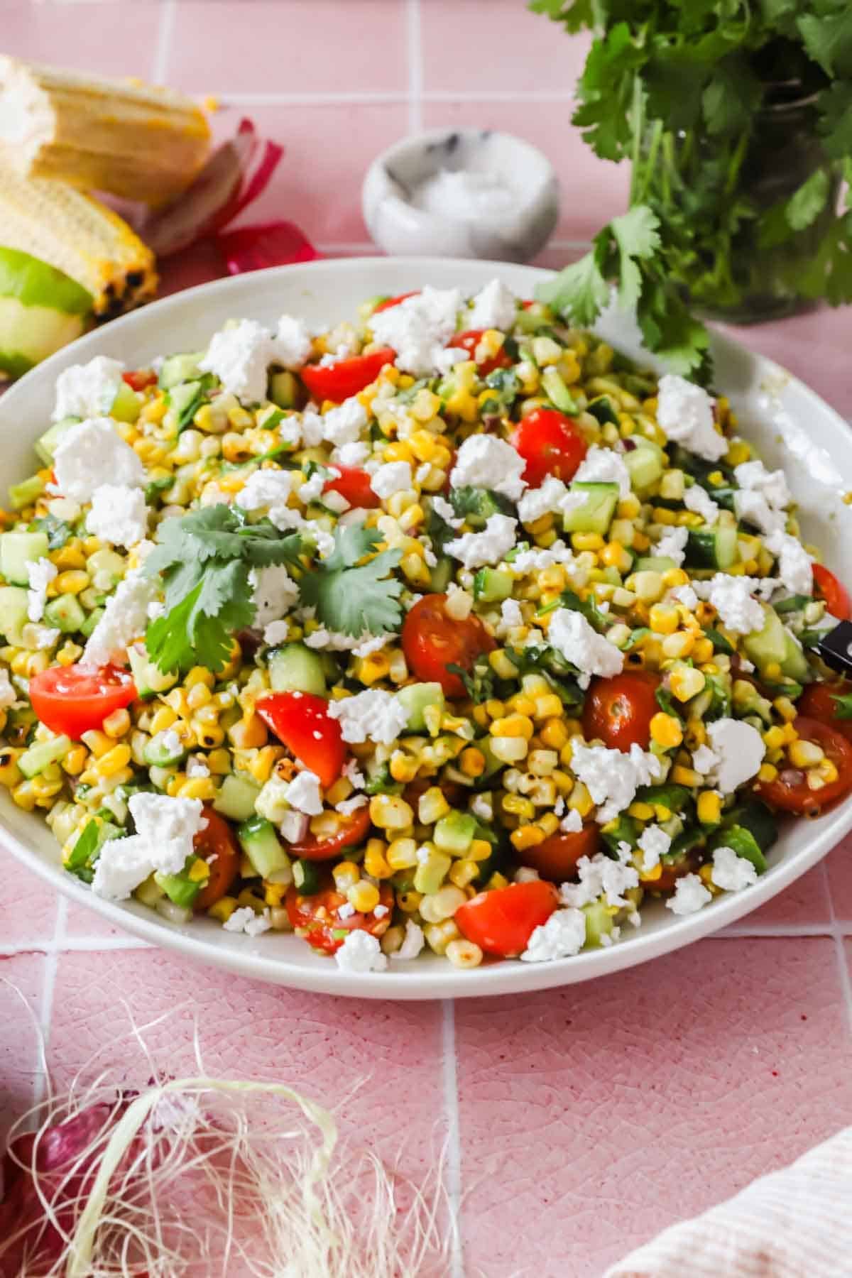 A plate of corn salad with cherry tomatoes, cucumber, cilantro, and crumbled cheese on a pink tiled surface.