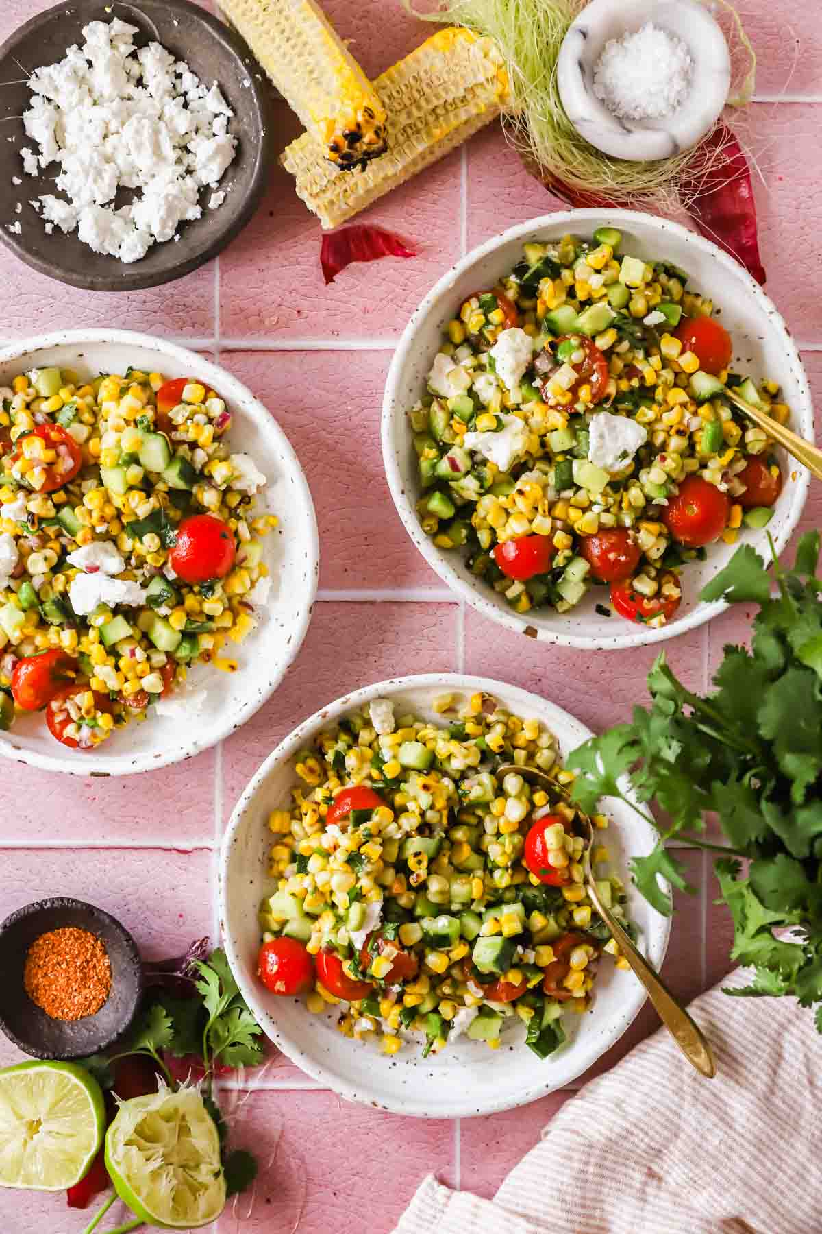 Three bowls of corn salad with cherry tomatoes, cucumber, and feta cheese, surrounded by corn cobs, lime halves, cilantro, seasonings, and a bowl of crumbled cheese on a pink tiled surface.