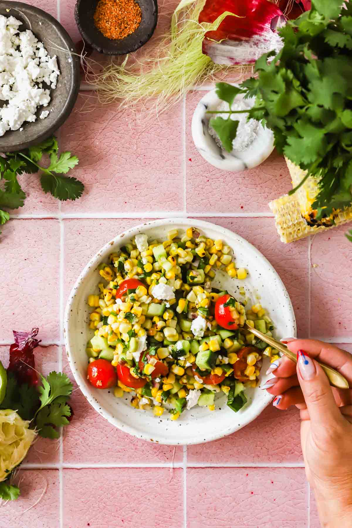 A hand holds a fork over a bowl of salad with corn, tomatoes, cucumber, feta, and herbs on a pink tiled surface, surrounded by fresh ingredients.