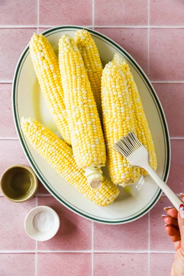 Five ears of corn on a plate, with a hand brushing one ear; a small bowl of oil and a bowl of salt are placed beside the plate on a pink tiled surface.