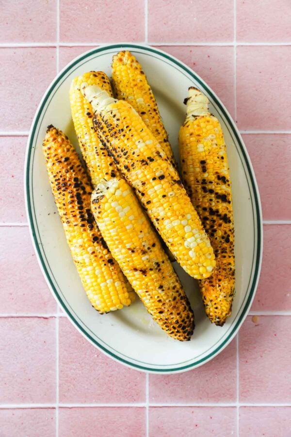 Six grilled corn cobs arranged on a white oval plate, set on a pink tiled surface.