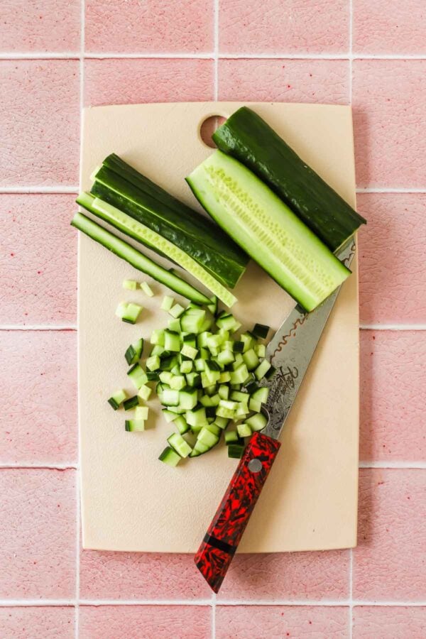 Diced, sliced, and whole cucumber pieces with a knife on a white cutting board, placed on a pink tiled surface.